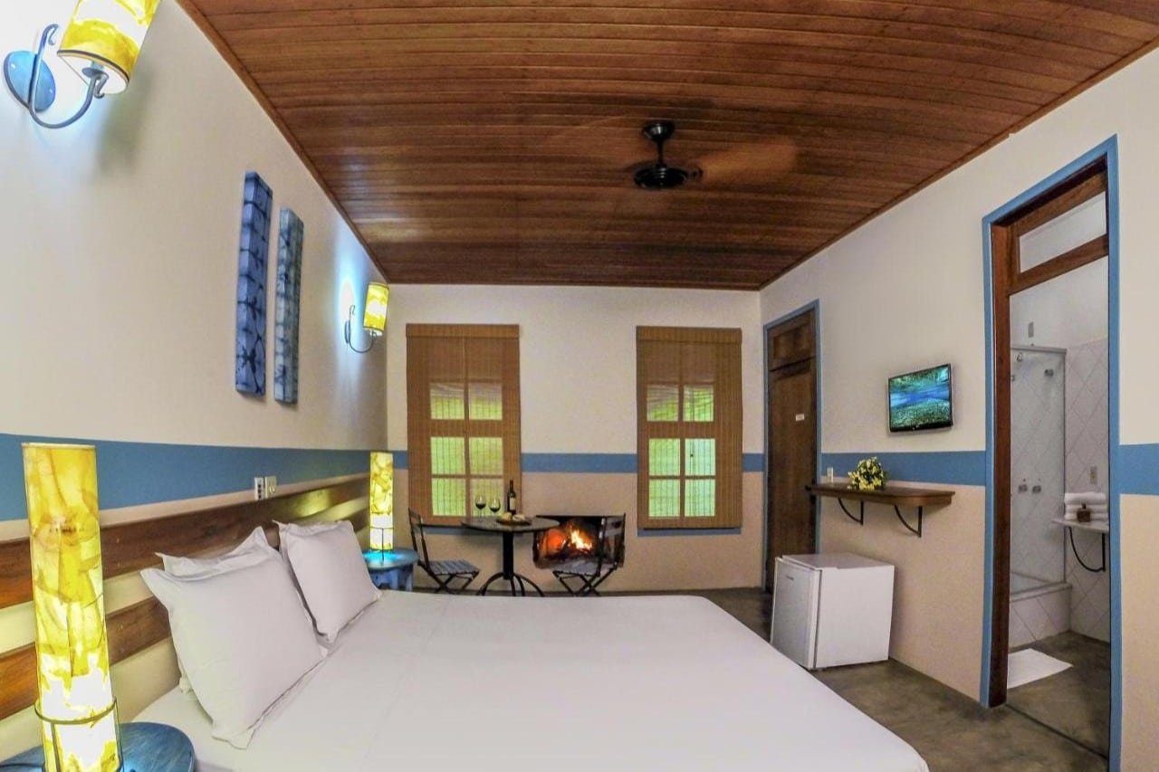 Um quarto aconchegante da Pousada Nascentes da Fortaleza, com uma cama grande, arandelas, teto de madeira, lareira pequena, frigobar e porta do banheiro à direita.