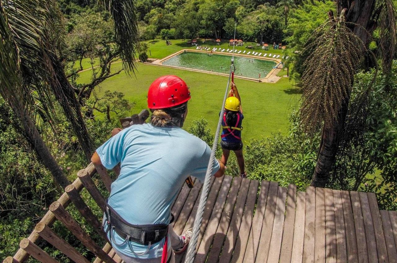 Duas pessoas usando capacetes e cintos de segurança estão em uma tirolesa com uma piscina e cadeiras de banho de sol visíveis ao fundo. O local fica na Pousada Nascentes da Fortaleza, um dos hotéis fazenda em Minas Gerais.