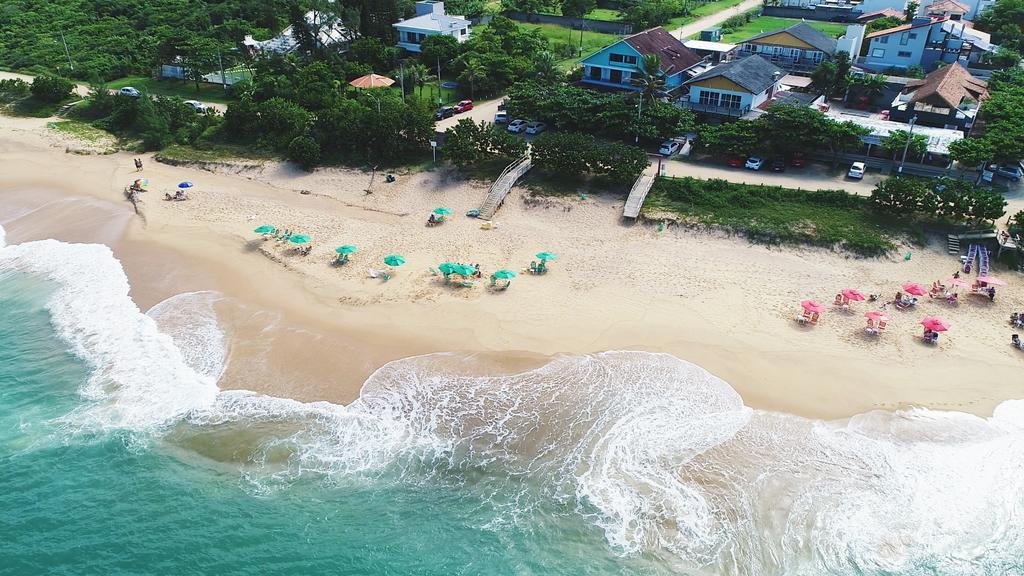 Casas localizadas à beira-mar, sendo uma delas a Pousada OceAnas Frente Mar Estaleiro, em Balneário Camboriú. Há bastante vegetação no arredor das casas e alguns corredores de madeira dando acesso à praia, que tem alguns guarda-sóis na areia bege e o mar tem um tom esverdeado