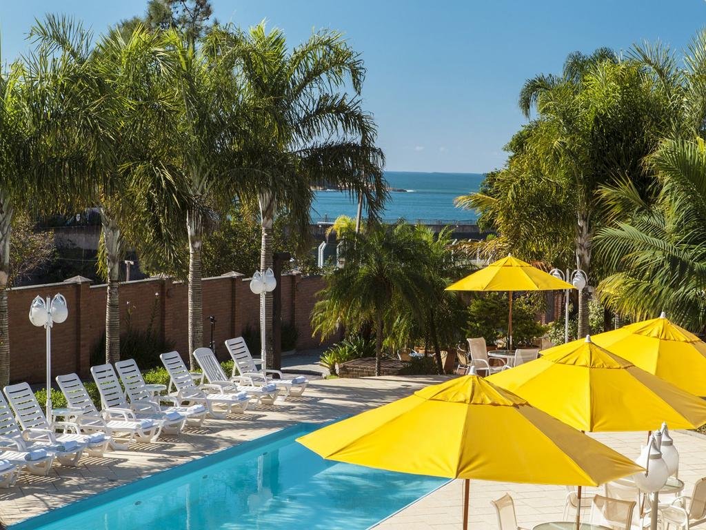 Piscina da Pousada Ponta do Lobo, com espreguiçadeiras brancas enfileiradas no lado esquerdo e, no direito, há guarda-sóis amarelos. O espaço tem bastante árvores e no fundo da imagem há vista para o mar