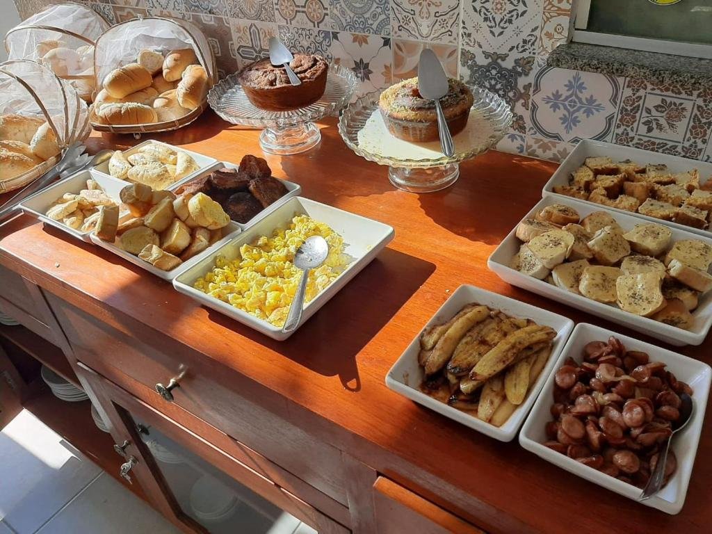 Café da manhã na Pousada Praia Bonita em Milagres, com tigelas cheias de quitutes diversos, como pães torrados, linguiça calabresa, banana frita, ovo mexido, e há ainda dois bolos e cestos com pães frescos