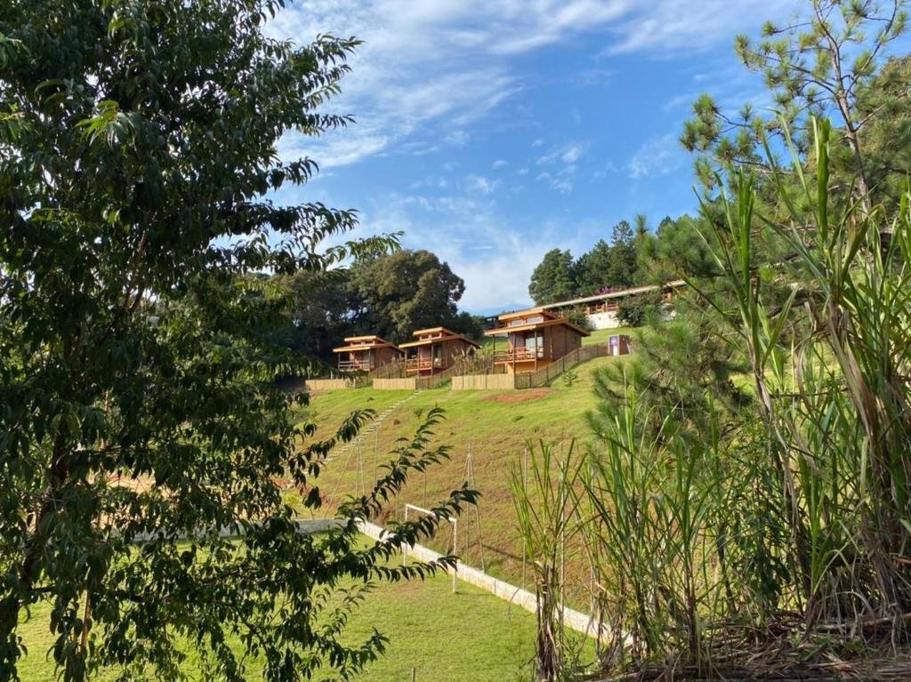 Casas em uma colina gramada cercada por árvores, sob um céu azul com nuvens dispersas no Pousada Recanto Primavera, um dos hotéis fazenda em São Paulo.