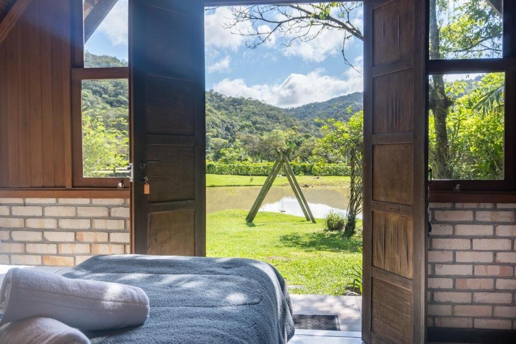 Interior de um dos chalés da Pousada Rio da Prata, de 20 m², com uma cama com duas toalhas brancas enroladas em cima e uma porta de madeira aberta mostrando a vista da natureza verde vasta localizada na área externa do local. Há também um lago refletindo o céu ensolarado