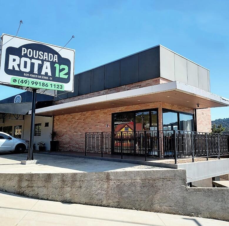 Fachada da Pousada Rota12, uma das pousadas na Serra do Rio do Rastro, com uma grande em placa em frente e carros estacionados ao lado esquerdo.