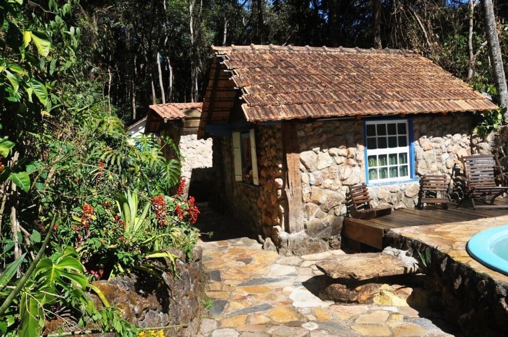 Chalé da Pousada Rural Lua Bonita do Neroaa com paredes feitas de pedras e uma piscina com cadeiras em volta no lado direito para ilustrar as pousadas em Macacos. Há bastante vegetação em volta.