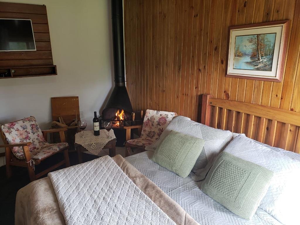 Quarto da Pousada Rural Vista Alegre, uma das pousadas na Serra do Rio do Rastro. Uma cama de casal está ao lado direito, e na lateral esquerda fica uma área de estar com TV, lareira, cadeiras e mesinha.