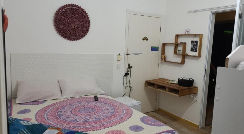 Quarto com uma colcha com estampa redonda, um móvel decorativo de parede, uma prateleira de canto com itens e um pequeno armário branco. Essa é a Pousada Shiva, uma das recomendações de pousadas em São Sebastião.