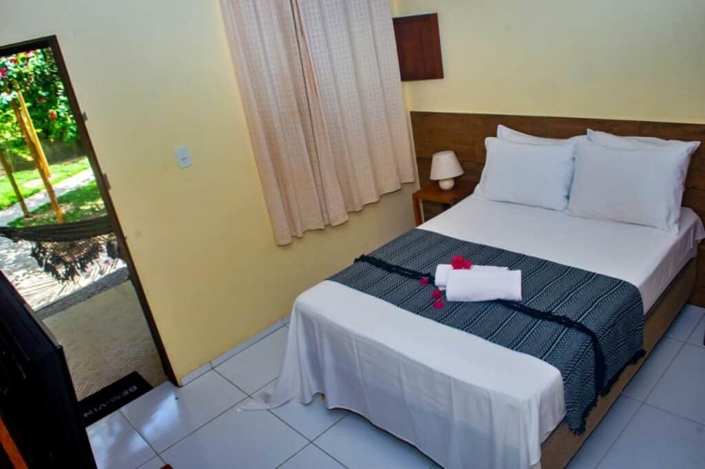 Quarto de casal na Pousada Sitio Araça, em São Miguel dos Milagres, com cama de casal, cortina na janela, duas toalhas enroladas sobre a peseira azul da cama, que está com lençóis brancos