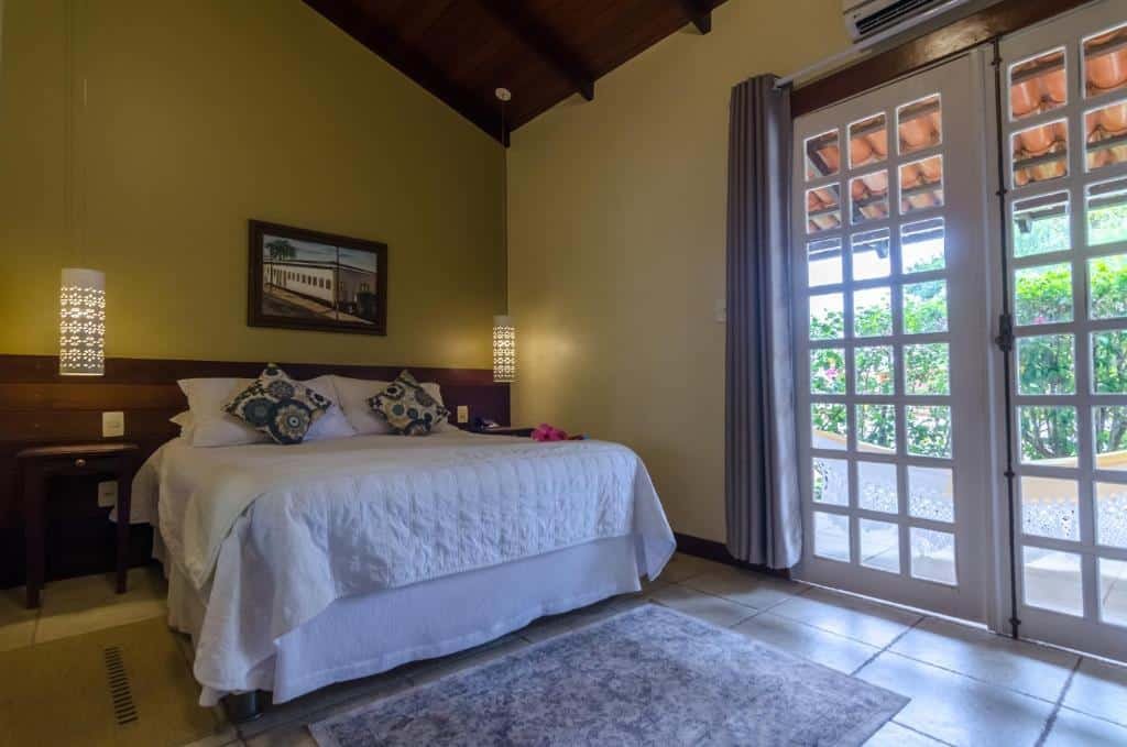 Quarto com cama de casal em Pousada Tajupá, coberto por edredom branco com travesseiros, mesa de cabeceira, quadro na parede e uma sacada com uma rede e vista para vegetação. 