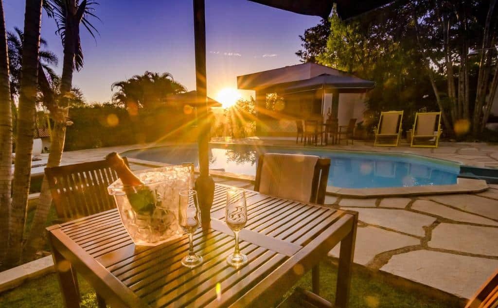 Área da piscina em Pousada Tajupá, com pôr do sol, área verde ao entorno e em primeiro plano duas taças de champagne em cima de um mesa de madeira. 