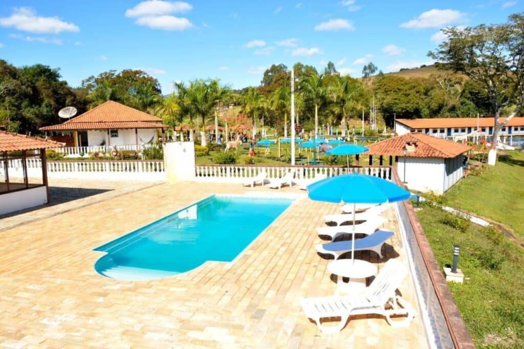 A imagem mostra uma piscina retangular ao ar livre, cercada por espreguiçadeiras brancas e guarda-sóis azuis em uma área pavimentada da Pousada temática Estrada Real. Ao fundo, há um gramado com palmeiras e estruturas de lazer, além de construções com telhados de telhas vermelhas, que parecem ser chalés ou áreas de descanso. O céu está claro, com poucas nuvens, e o ambiente é ensolarado, criando uma atmosfera agradável para relaxar. A paisagem ao redor é verde e inclui colinas e árvores, complementando o ambiente de tranquilidade.