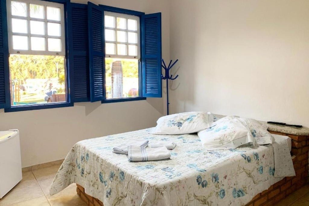 A imagem mostra um quarto iluminado com uma cama de casal coberta por uma colcha floral em tons de branco e azul da Pousada temática Estrada Real. A cama está posicionada ao lado de uma janela grande com moldura e venezianas azuis abertas, permitindo a entrada de luz natural. Ao lado da cama, há um pequeno frigobar branco, e na parede à direita, um cabideiro azul está fixado. O ambiente é simples, com paredes brancas e piso de cerâmica, transmitindo uma sensação de tranquilidade e conforto. A vista pela janela revela uma área externa com árvores e jardins.