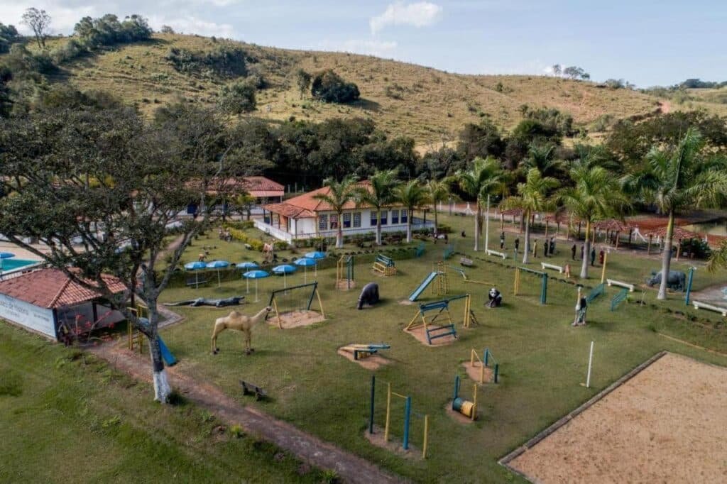 A imagem mostra um parque ao ar livre com diversas áreas de lazer e atividades. Há brinquedos de playground como balanços, escorregadores e outros equipamentos, espalhados em um amplo gramado. Estátuas de animais como camelo, urso e rinoceronte também decoram o local. Ao fundo, há uma construção com telhado de telhas vermelhas e paredes brancas, em estilo colonial. O parque é cercado por colinas e árvores, e algumas palmeiras estão dispostas ao longo da área. Vários guarda-sóis azuis fornecem sombra para as áreas de descanso. Essa é a Pousada temática Estrada Real, um dos hotéis-fazenda em Minas Gerais.