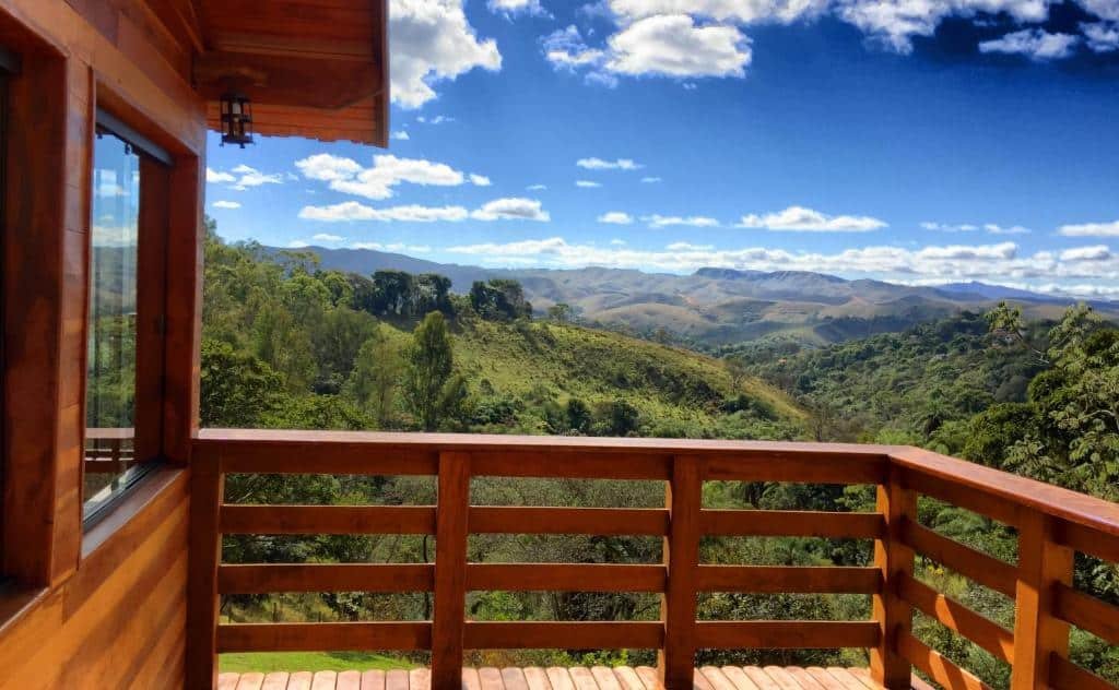 Varanda de um chalé com deck e paredes de madeira, com vista panorâmica da natureza, incluindo montanhas no horizonte, céu azul com nuvens brancas e bastante vegetação verde