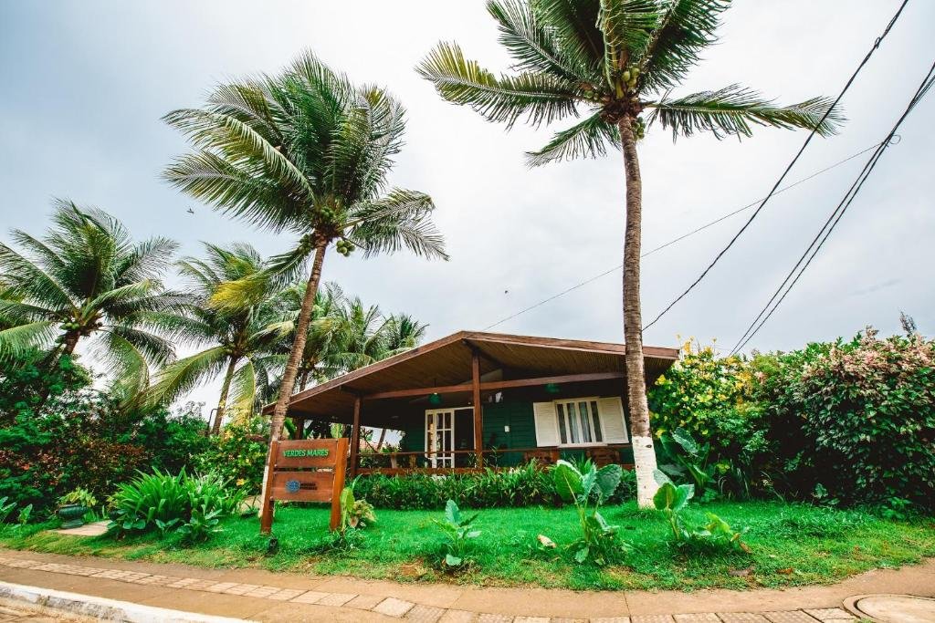 Fachada da Pousada Verdes Mares, em Fernando de Noronha, sendo a casa envolta por arbustos verdes, gramado em frente e dois coqueiros altos de cada lado