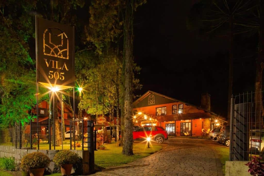 A imagem mostra uma cena noturna de uma propriedade com uma placa iluminada na entrada que diz “VILA 505”. Há uma entrada de paralelepípedos que leva a um edifício que parece ser um restaurante ou pousada, com iluminação quente e várias janelas brilhando na escuridão. Árvores e folhagens são visíveis ao redor da propriedade, e há um portão aberto à esquerda da placa.