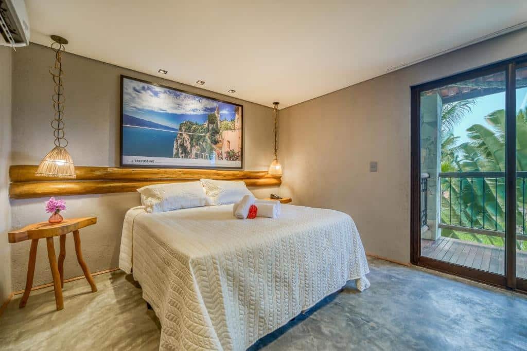 Quarto de casal na Pousada Villa Italiana, em São Miguel dos Milagres, com cama alta e colcha creme, paredes cor de areia, um quadro com destino de corta, luzes pendentes uma em cada lado, sobre a mesinha de cabeceira, e uma sacada