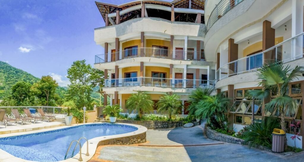 Área externa do Hospedaria Vitória Mar. Do lado direito e no fundo a varanda dos quartos e o jardim. Do lado esquerdo uma piscina e cadeiras de tomar sol.