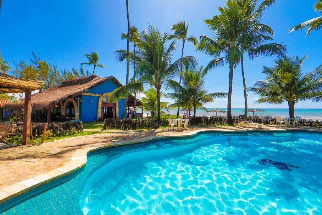 Piscina da Pousada Xalés de Maracaípe com água azul cristalina cercada por palmeiras e um edifício com telhado de palha sob um céu ensolarado.