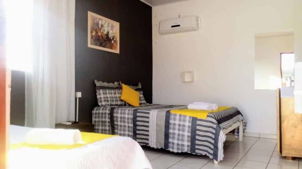 Camas em quarto da Pousada Zoe em Pirenópolis, cama coberta com edredom de estampa com travesseiros, quadro pendurado na parede, uma janela com cortina ao centro e frigobar em frente a uma das camas do quarto.