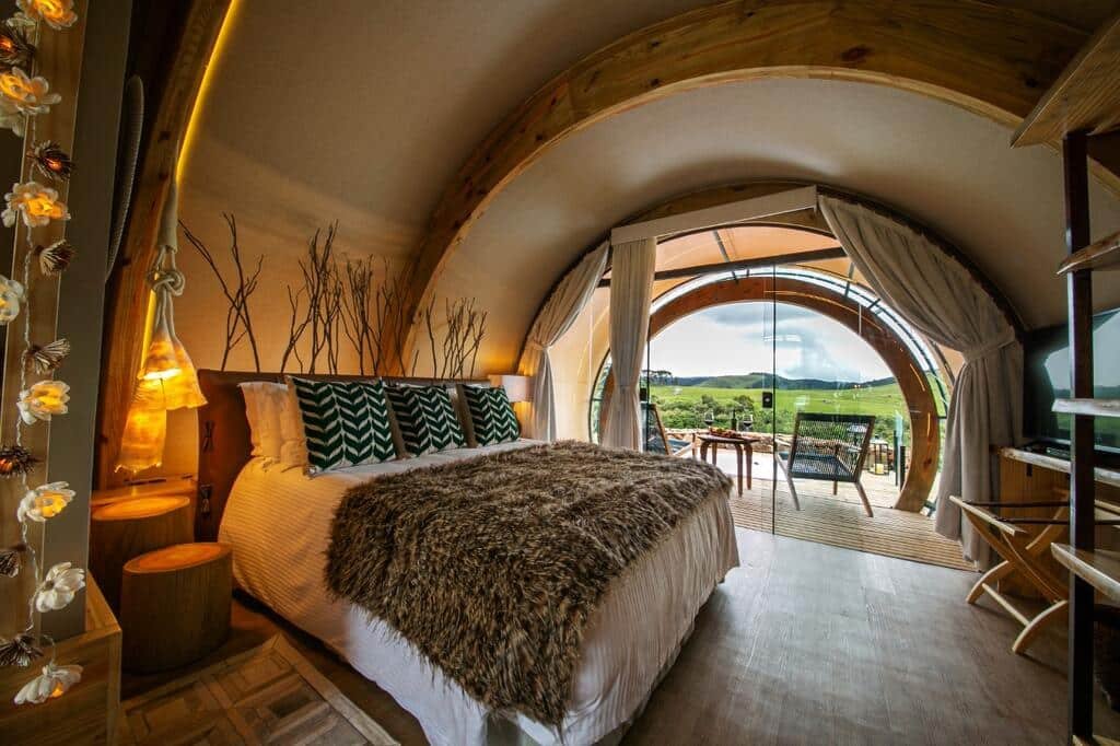 quarto do hotel Parador Cambará do Sul com vista para uma cama de casal com decoração rústica, arquitetura arredondada, uma área de estar com duas cadeiras e uma mesa pequena e redonda com vista para a paisagem