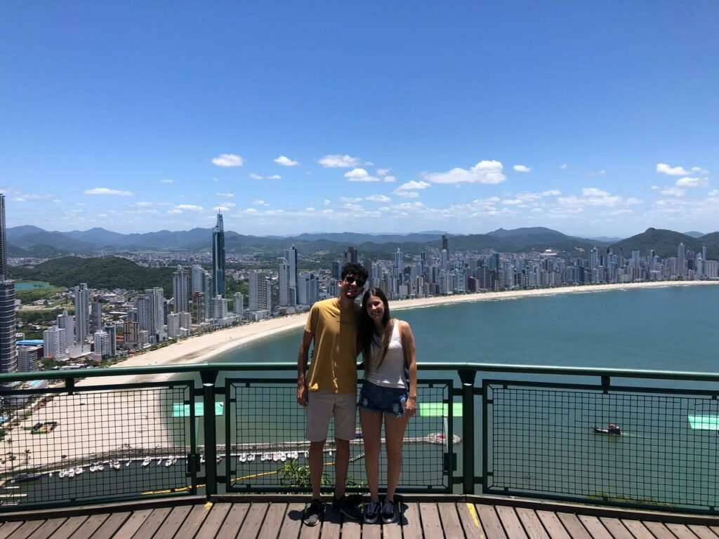 Um casal na frente, atrás uma grade de apoio, no fundo a vista da famosa Praia Central de Balneário Camboriú.