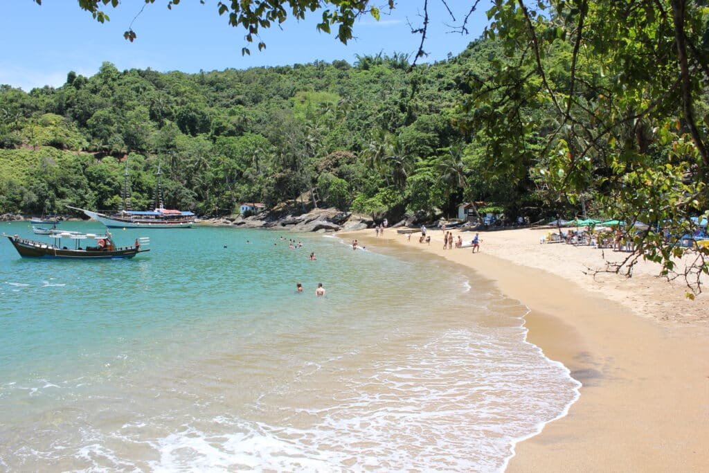 Praia de areia com pessoas nadando e tomando sol, um barco na água e árvores verdejantes ao fundo. Essa é a Praia da Fome em Ilhabela, no Litoral Norte de São Paulo.
