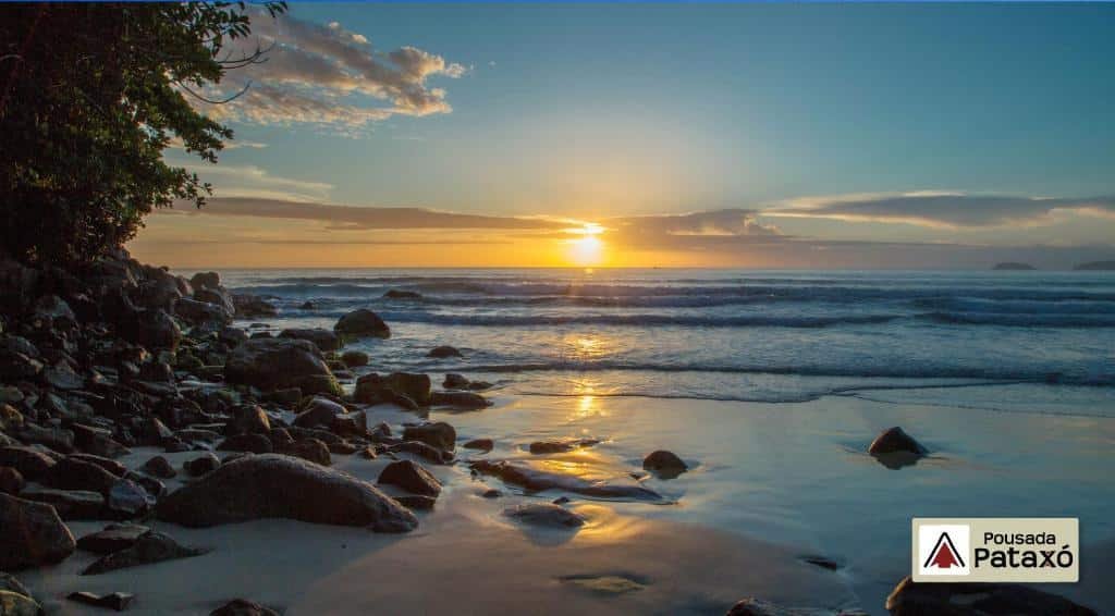 Praia das Toninhas em Ubatuba num fim de tarde com o sol de pondo, muitas pedras perto da areia e vegetação