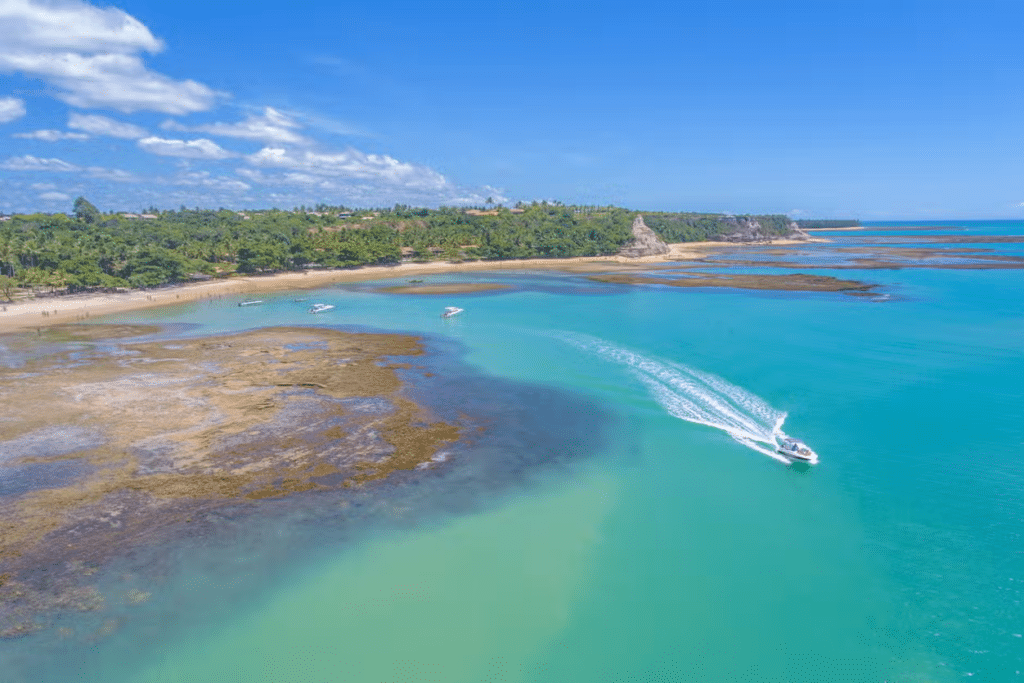 Vista aérea de uma lancha em águas azul-turquesa perto da Praia do Espelho com vegetação verde e céu azul claro acima. Imagem para ilustrar post sobre o que fazer no feriado de Corpus Christi.