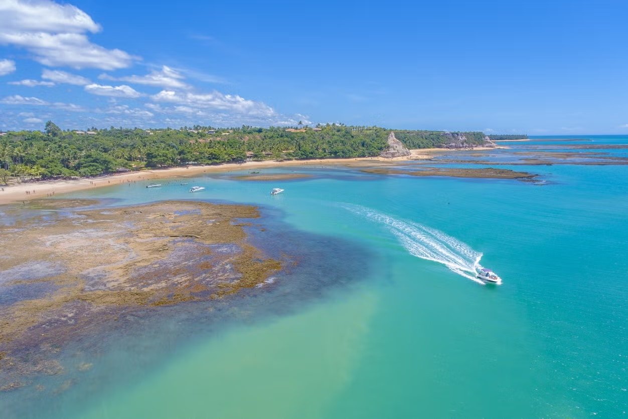 Uma foto aérea e panorâmica da Praia do Espelho, entre Trancoso e Caraíva. Mostra a água azul cristalina, com uma lancha passando, além do resto da geografia que compõe o lugar. Há árvores ao fundo. O céu é azul e com poucas nuvens. A foto representa o que fazer em Porto Seguro.