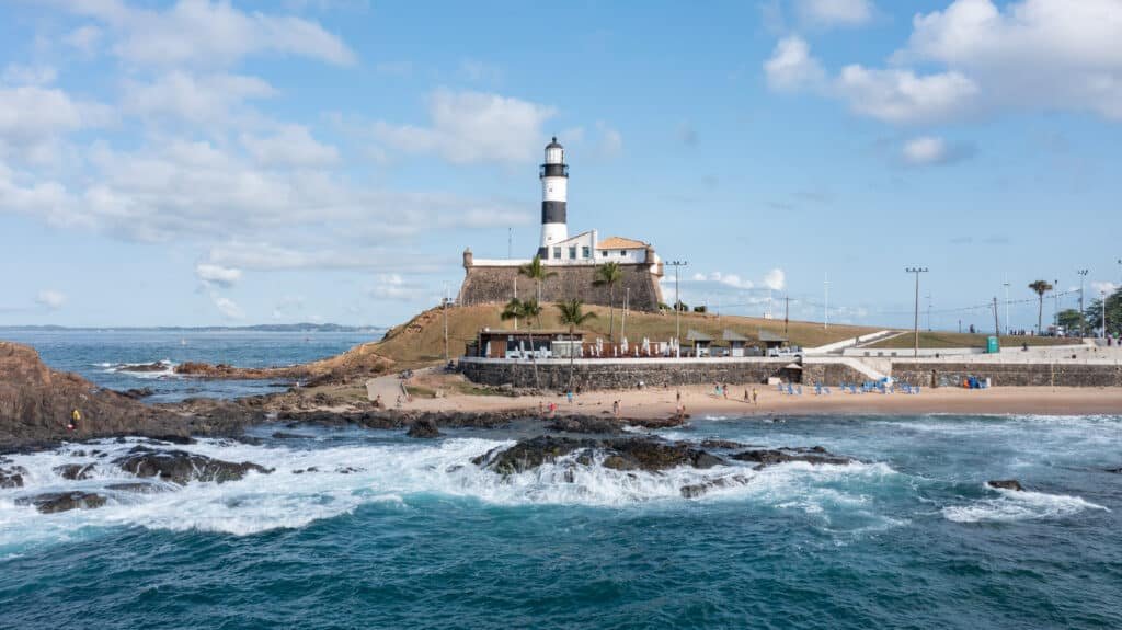 O Farol da Barra em uma colina tem vista para um litoral rochoso com uma pequena praia. Pessoas estão caminhando na areia sob um céu parcialmente nublado.