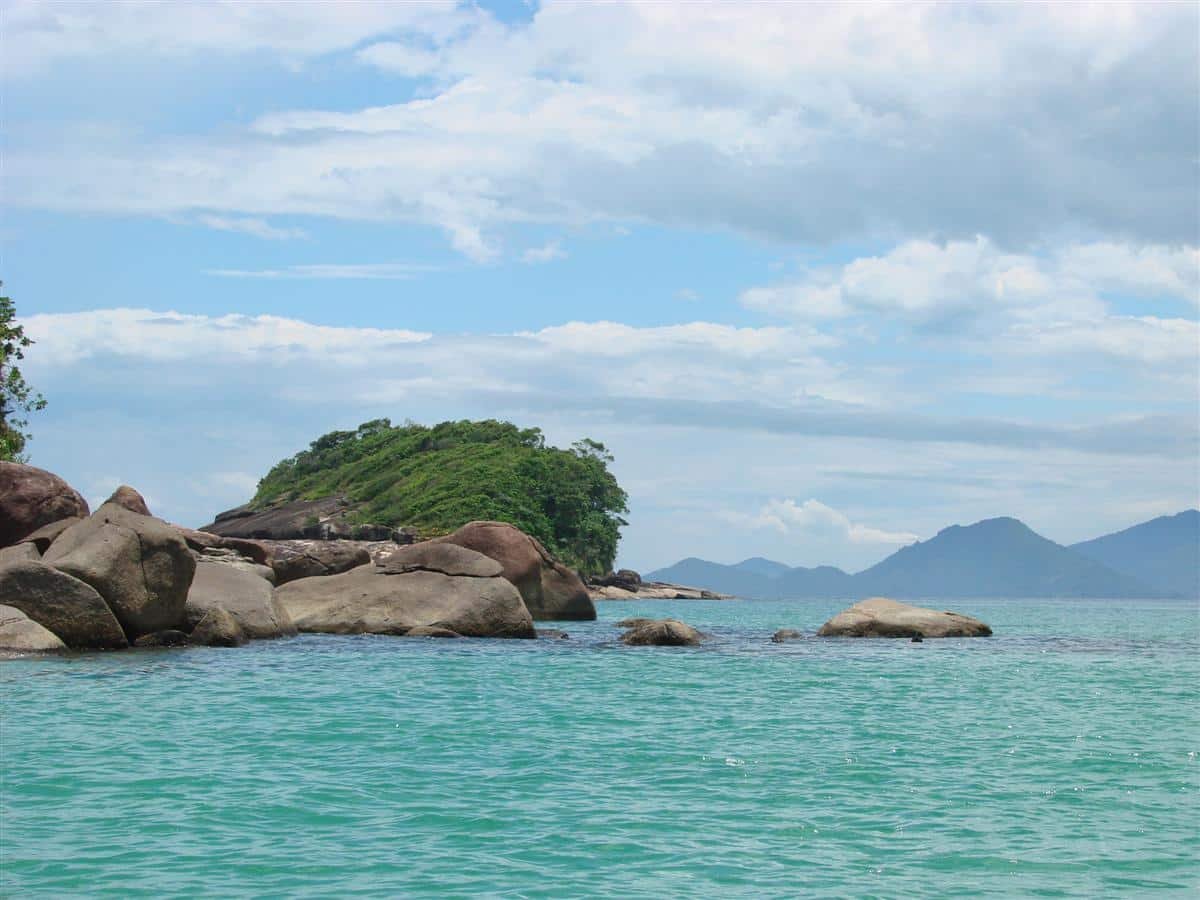 Praia de Perequê Mirim. O mar verde claro na frente, pedras no meio, e no fundo uma ilha e montanhas.