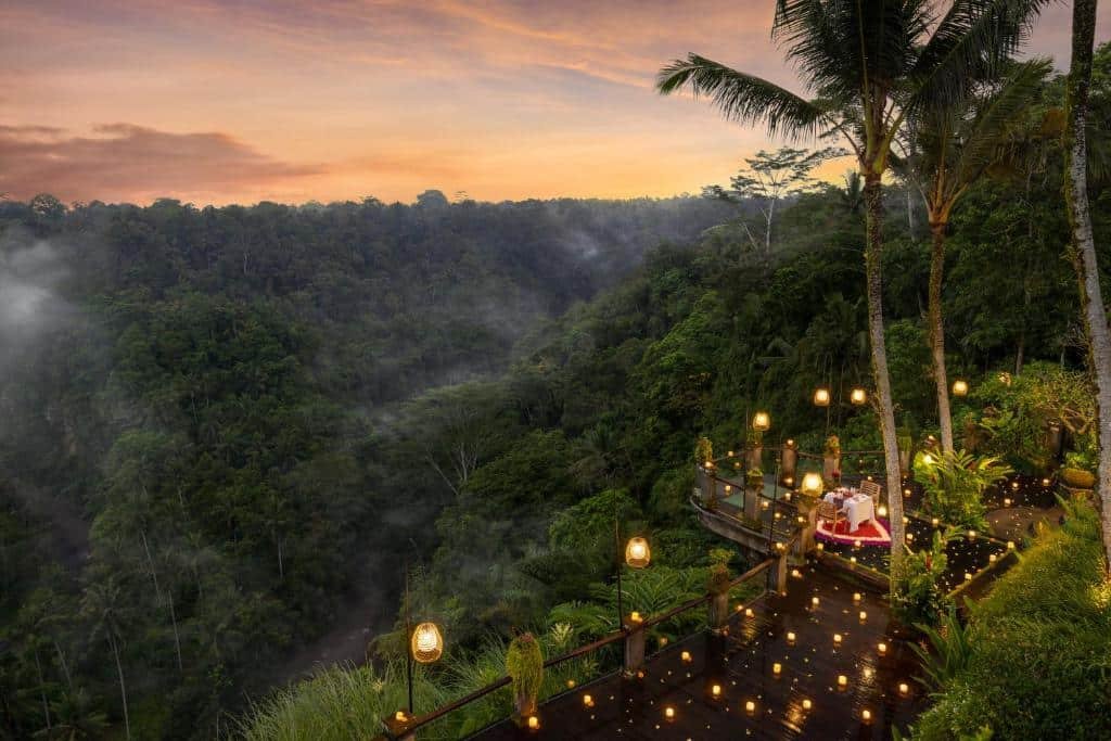 Uma paisagem de floresta tropical ao pôr do sol, com um deck de madeira iluminado, palmeiras e colinas cobertas de vegetação densa.