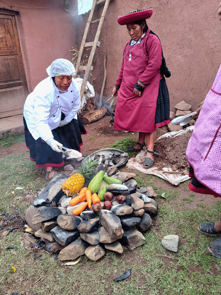 uma mulher peruana usando um avental e uma touca higiênica nos cabelos está inclinada sobre uma pilha de pedras no chão, onde se encontram alguns legumes e frutas que estão defumando. Ao lado desta mulher, há outra mulher auxiliando.