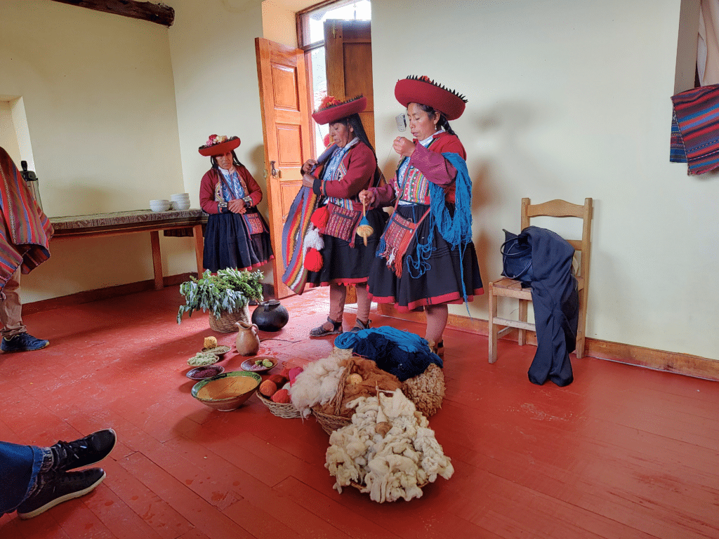 três mulheres peruanas vestidas tipicamente apresentando como se faz a tintura das cores das lãs. Elas estão em pé, atrás de vários objetos no chã, como lãs, tigelas de água e demais materiais.