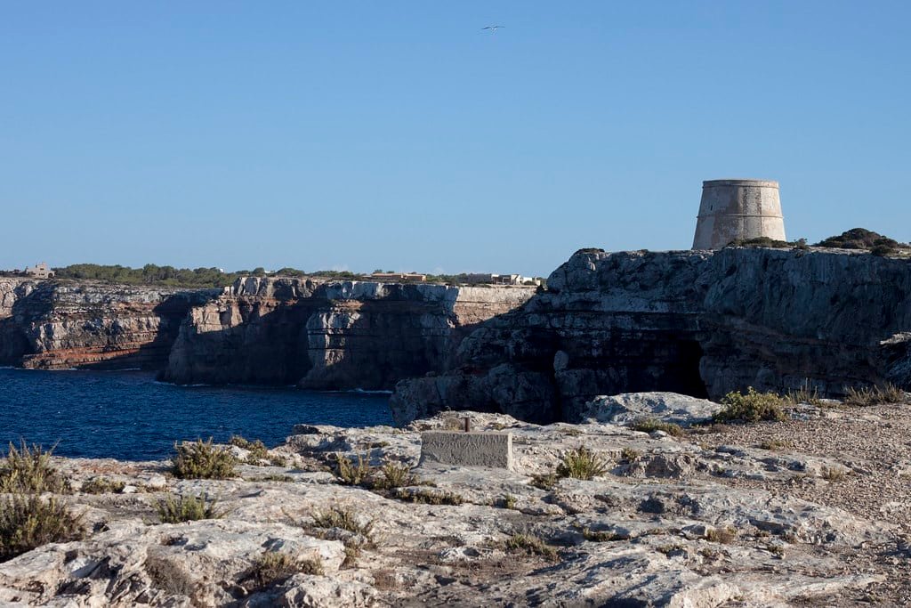 Paisagem costeira rochosa com uma torre de pedra nos penhascos, com vista para o mar sob um céu azul claro. Vegetação esparsa em primeiro plano. Essa é a Torre de Punta Prima, uma das opções de o que fazer em Formentera.