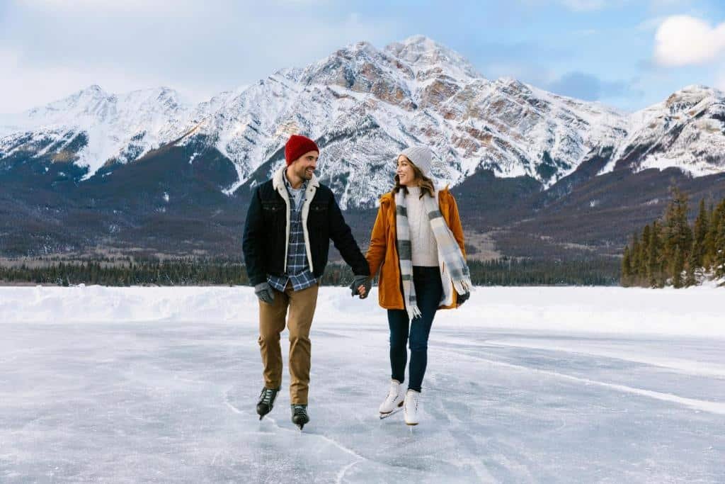 Um homem e uma mulher patinando de mãos dadas em um lago congelado com montanhas cobertas de neve ao fundo para representar um dos melhores lugares para lua de mel