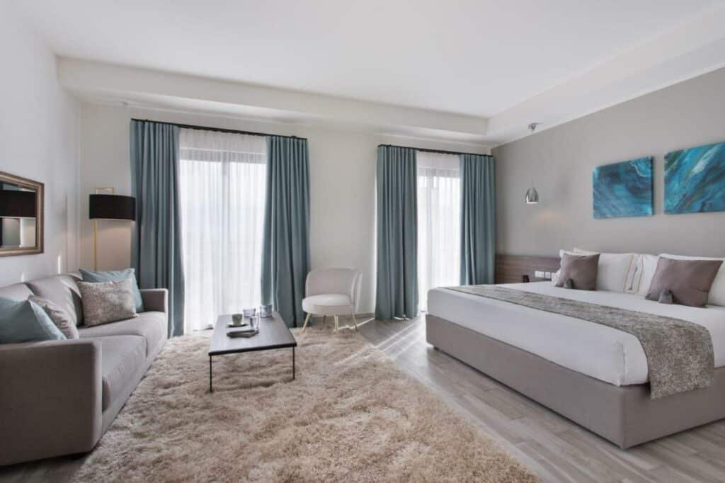 Quarto espaçoso no 1926 Hotel & SPA com duas sacadas, um sofá cinza com almofadas azuis, uma tapete bege, uma mesinha de centro em madeira, uma poltrona branca e uma cama de casal nos mesmos tons de cores, na parede há dois quadros azuis sob a cama, para representar hotéis em Malta