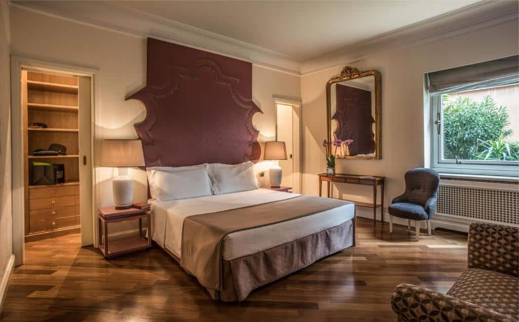 quarto do 47Luxury Suites - Colosseo, uma das opções de onde ficar em Roma, com cama de casal, mesinha e luminária de cabaceira de ambos os lados, espelho grande com moldura trabalhada, mesa e cadeira