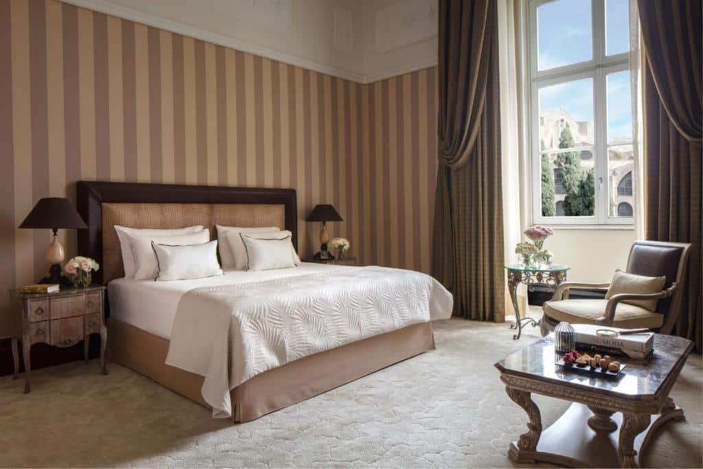 quarto do Anantara Palazzo Naiadi Rome Hotel, um dos hotéis de luxo em Roma, com cama de casal bem grande, mesinha e luminária de ambos os lados, mesa de centro em madeira com poltronas, detalhes brancos e vista da cidade pela janela grande 