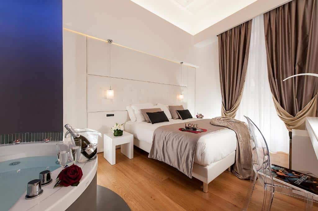quarto do Argentina Residenza Style Hotel, um dos hotéis no centro de Roma, com cama de casal grande, mesinha com luminárias compridas de ambos os lados, banheira de hidro do outro lado com champanhe, e uma grande janela com cortinas pesadas, à frente, mesa com cadeira transparente