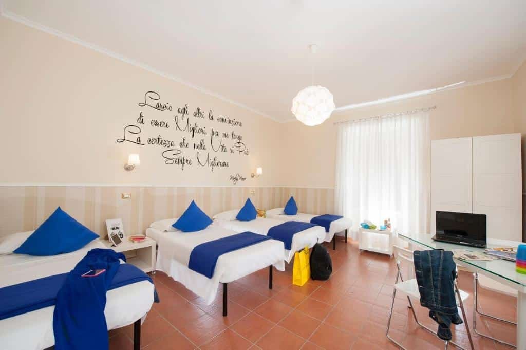 quarto do Budget Rooms Des Artistes, um dos hostels em Roma, com várias camas de solteiro lado a lado, janela grande com cortinas, escritos na parede e mesa com cadeiras transparente, os detalhes são em azul e o guarda-roupa é branco