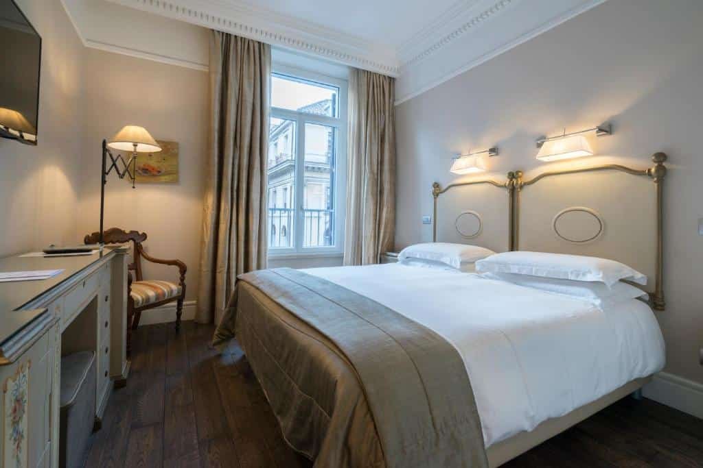 quarto do Casa Montani, um dos hotéis boutique em Roma, com cama de casal, luminárias acima da cabeceira e, à frente uma bancada com mesa e tv, do outro há uma poltrona e janela com cortinas, tudo em tons de marrom claro e branco