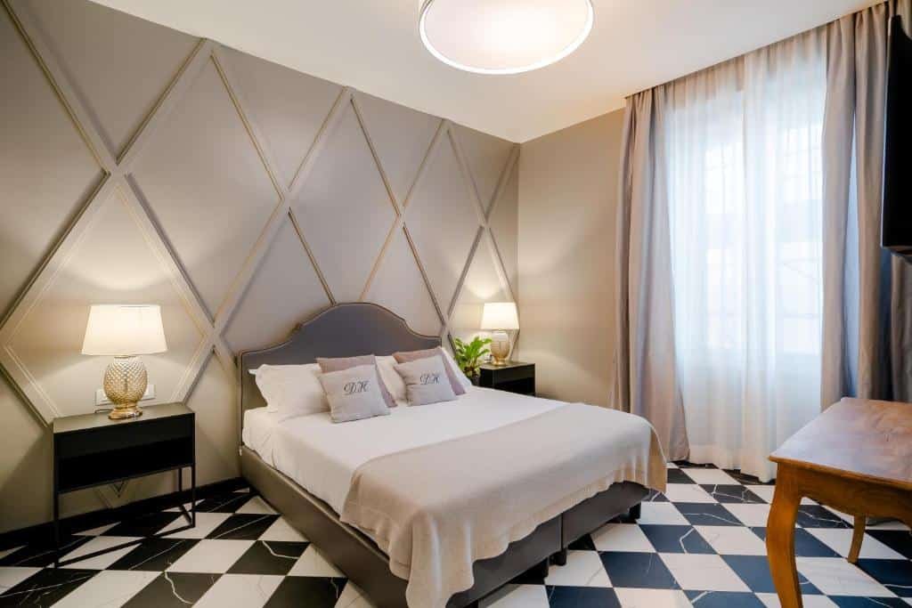 quarto do Deseo Home, um dos hotéis boutique em Roma, com cama de casal grande, mesa com luminária de ambos os lados, papel de parede trabalhado atrás, chão xadrez e janela grande com cortinas claras, tudo em tons de bege