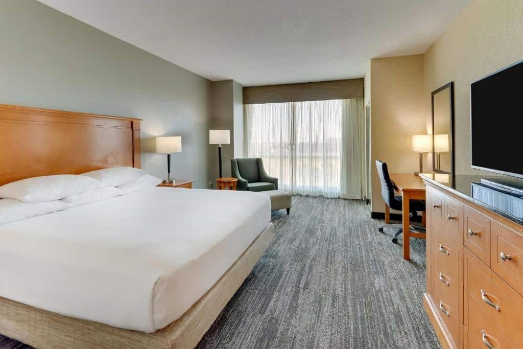 quarto amplo do Drury Inn & Suites Orlando near Universal Orlando Resort com mesa e cadeira, cômoda com tv em cima, poltrona em frente a uma janela de vidro com cortinas e uma cama com luminárias e mesinhas de ambos os lados