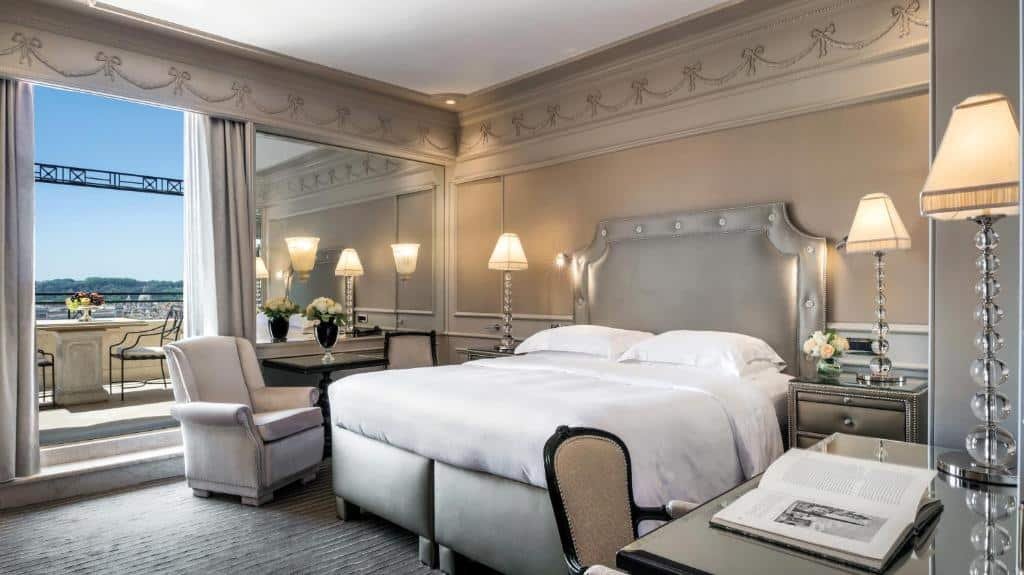 quarto do Hassler Roma, um dos hotéis de luxo em Roma, com cama de casal bem grande, mesinha e luminária de ambos os lados, mesa com poltronas, de um lado com sacada e vista e, do outro, mesa com cadeiras, tudo com decoração clássica e clara