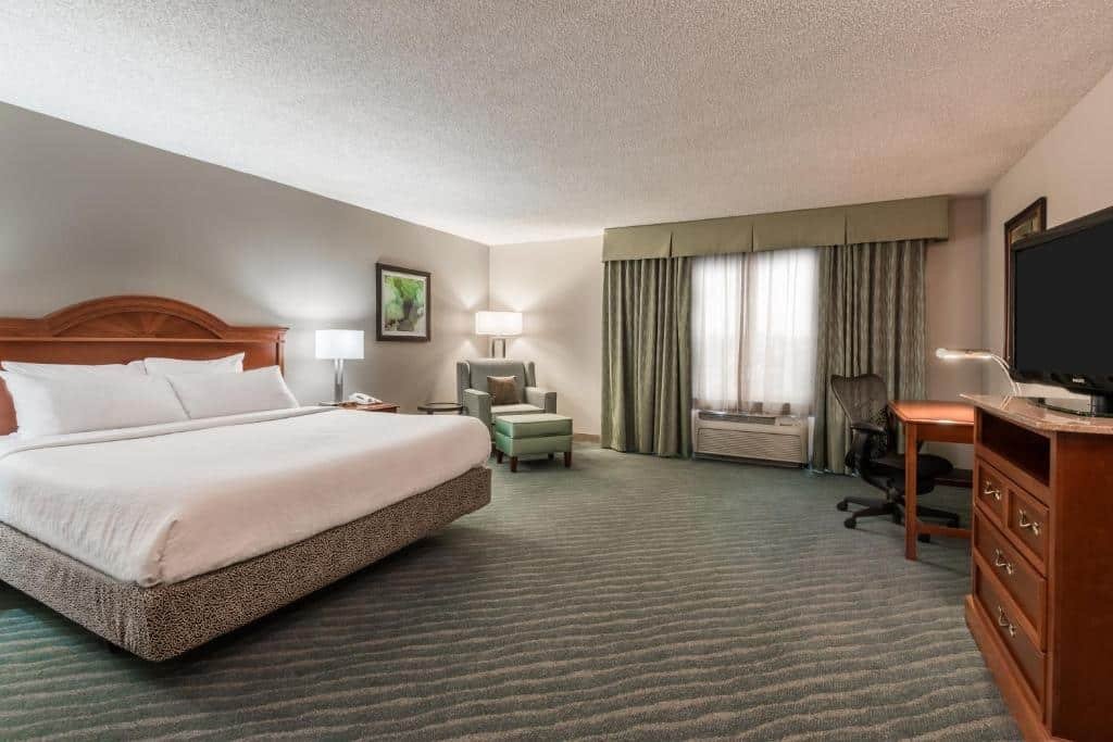 quarto amplo do Hilton Garden Inn Orlando International Drive North com janela grande com cortinas, cadeira e mesa, sofá e cama de casal com luminárias e mesinhas em ambos os lados no Hilton Garden Inn Orlando International Drive North