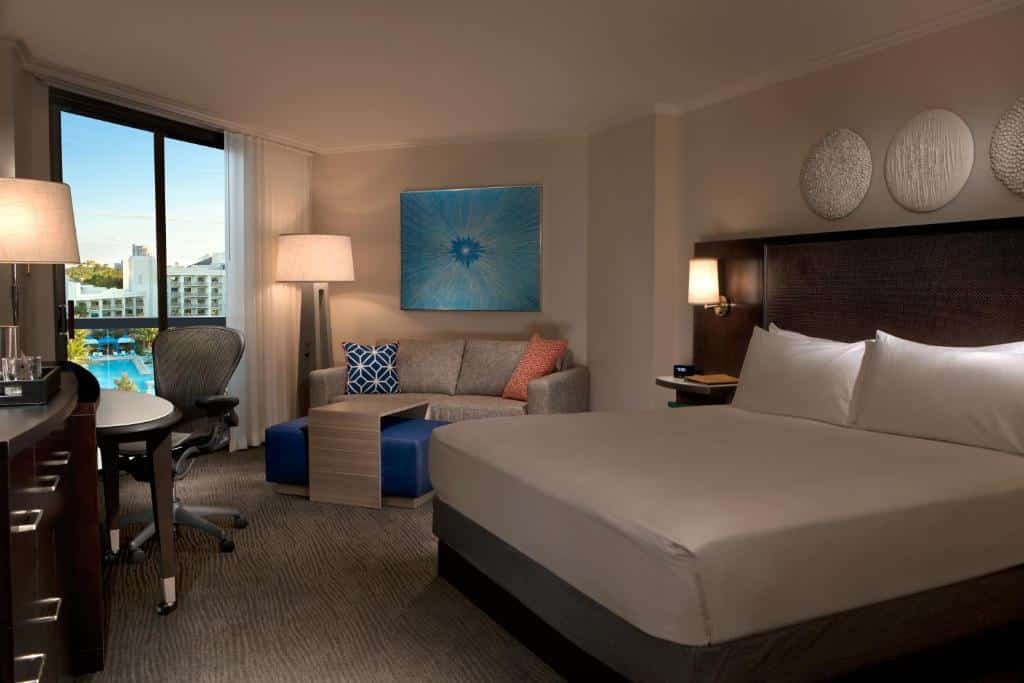 quarto do Hilton Orlando Buena Vista Palace com vista da piscina em janelas grandes com luminária de chão, há ainda um pequeno sofá, cômoda e tv em frente à cama com mesinha e luminária de ambos os lados