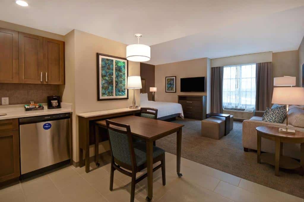 quarto amplo e completo, com cama, tv, sofá, mesa com cadeiras e cozinha com luminárias acima e tons de bege e verde no Homewood Suites By Hilton Orlando Flamingo Crossings, Fl