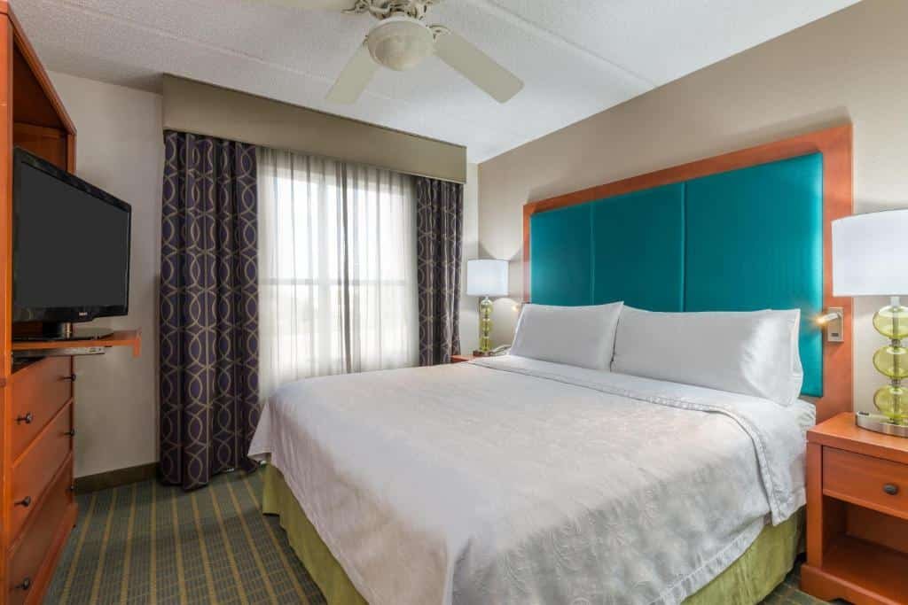 quarto com cama de casal branco com detalhes de cabeceira em verde, janela grande com vidro e cortinas escuras, tv dentro do guarda-roupa com uma estrutura giratória no Homewood Suites by Hilton Orlando-Nearest to Universal Studios